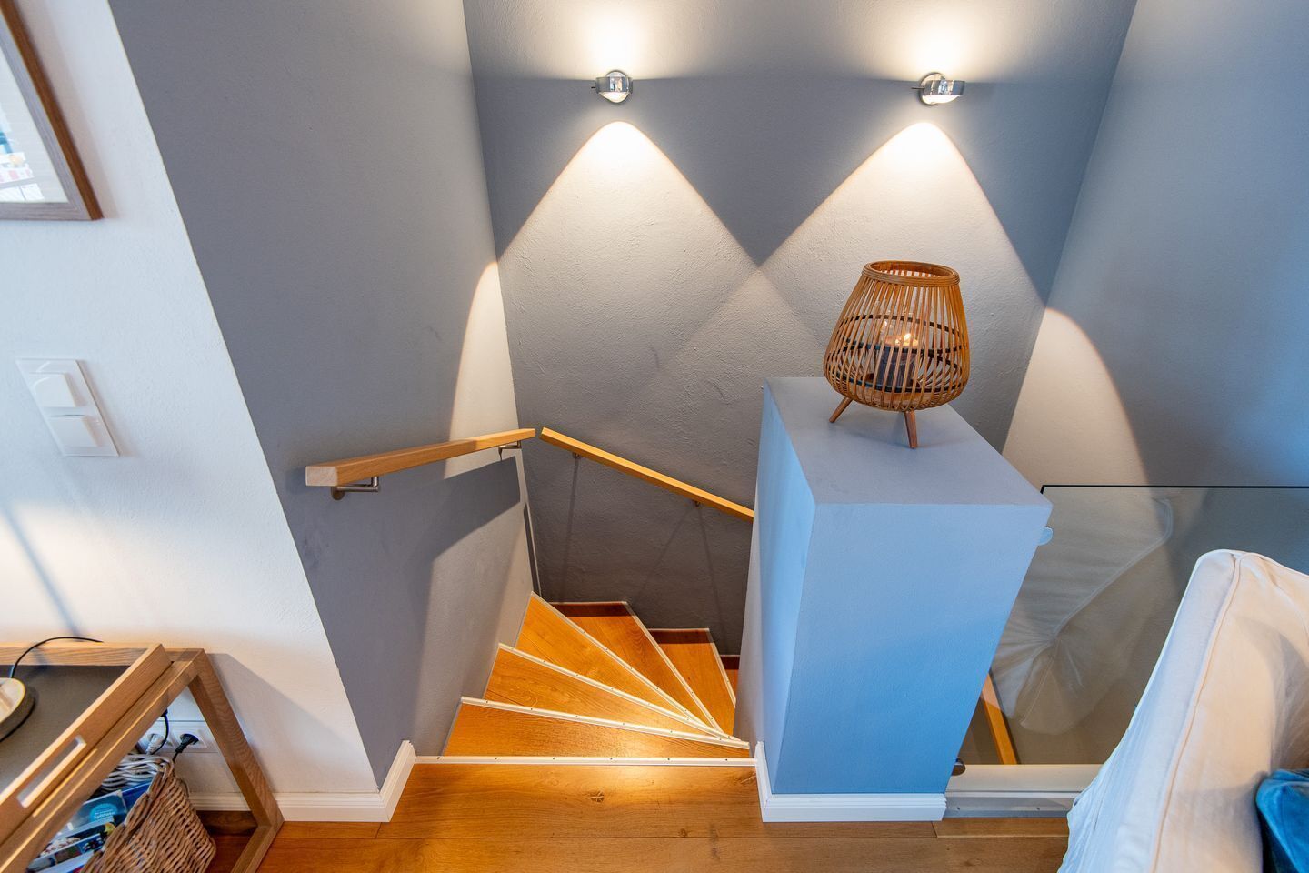 Una escalera de madera con barandilla que lleva a un rellano con una lámpara de mimbre y un banco acolchado blanco.