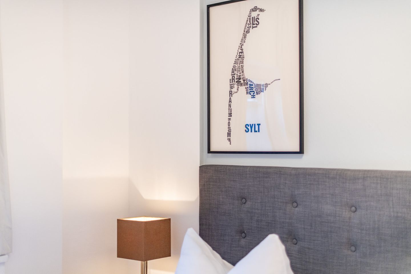 Ein modernes Zimmer mit grauem Stoffkopfteil, einem gerahmten abstrakten Kunstwerk an der Wand, einer Stehlampe und einem weißen Kopfkissen auf dem Bett.