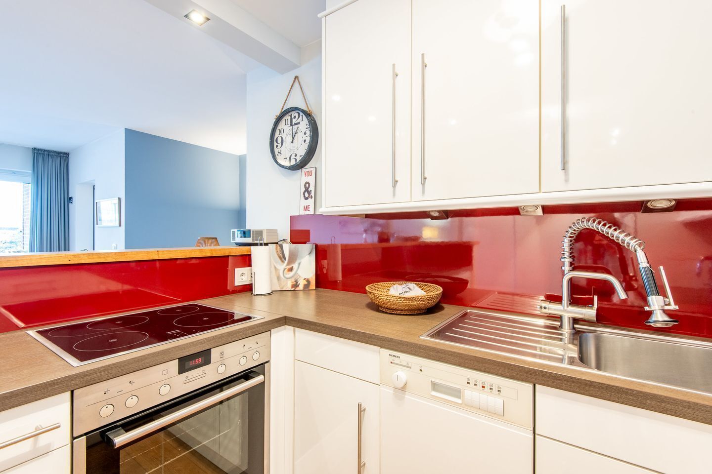 A modern kitchen with a red countertop, stainless steel appliances, white cabinets, and a wall-mounted clock.
