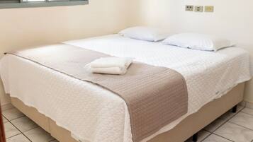 A bed with a beige frame and white bedding, a window with blinds, and a wall-mounted light fixture.