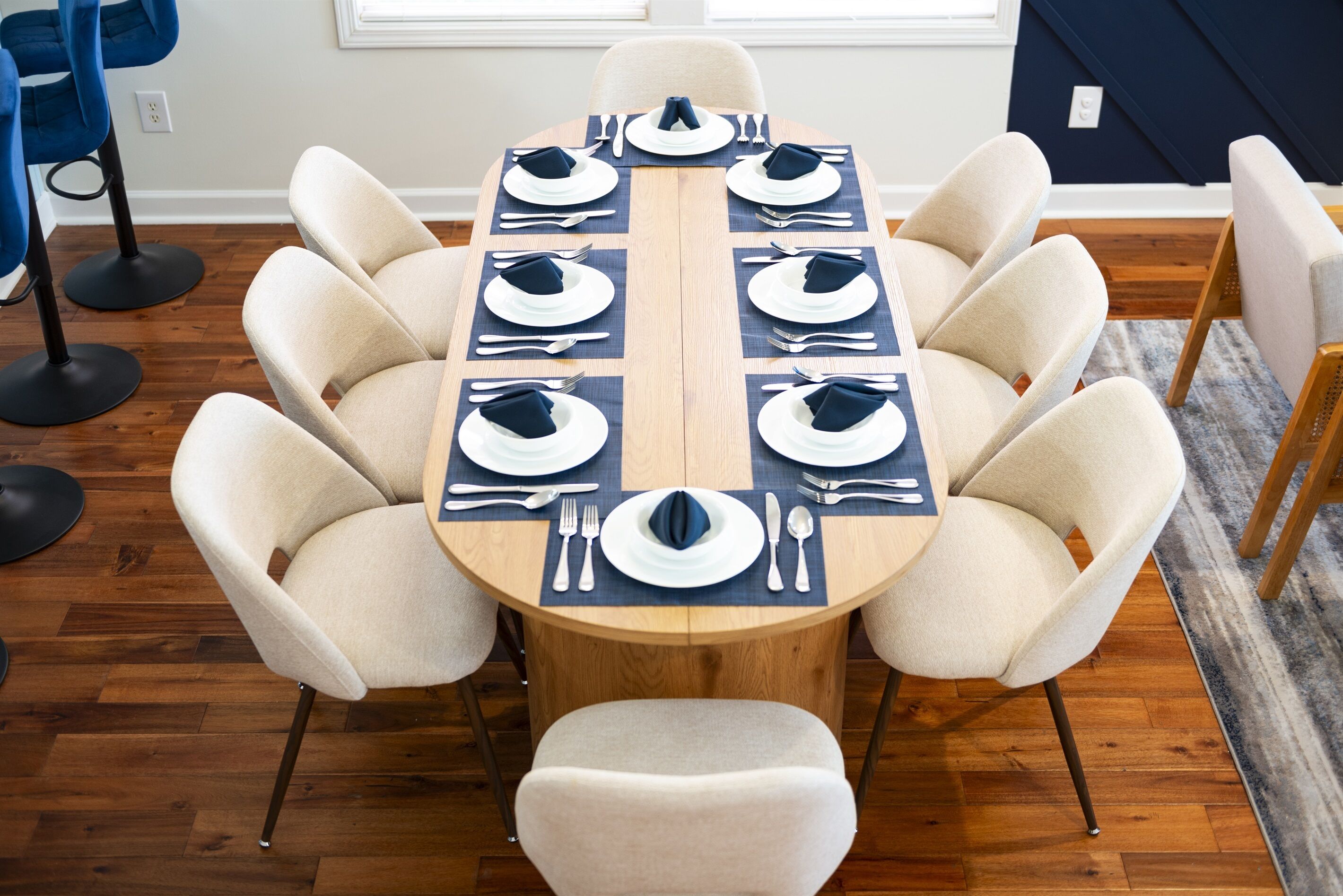 A round dining table with eight beige chairs, set with white plates, blue napkins, and silverware.
