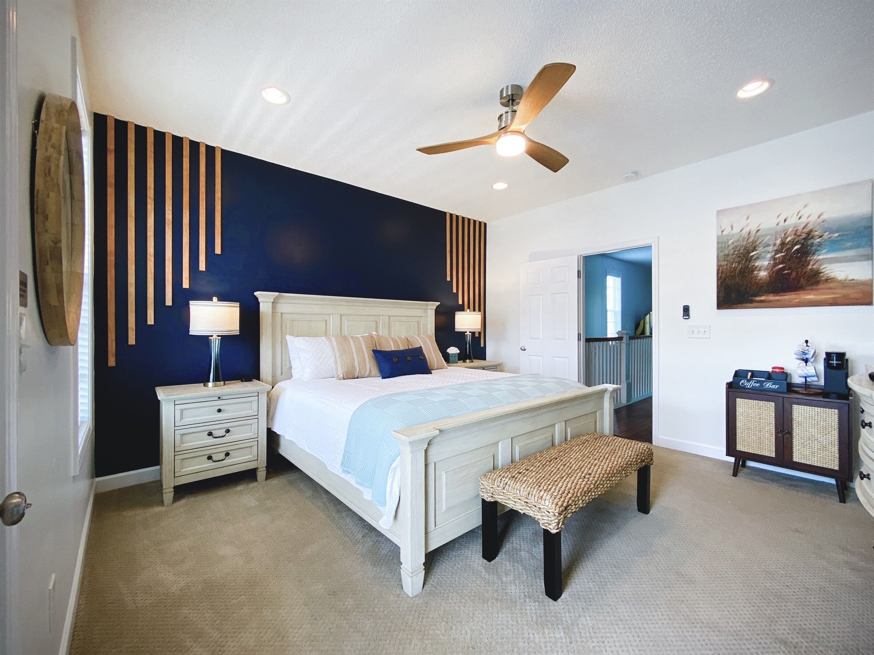 A bedroom with a bed, bedside tables, a ceiling fan, a bench, a coffee station, and a door leading to another room.