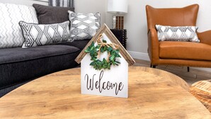 Une pancarte « Bienvenue » avec un cadre en forme de maison et une guirlande, posée sur une table en bois dans un salon comprenant un canapé et un fauteuil en cuir.