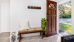 Un banc en bois avec une assise en osier, un coussin blanc et une plante en pot, placé à côté d’une porte en bois dotée d’une poignée en métal.