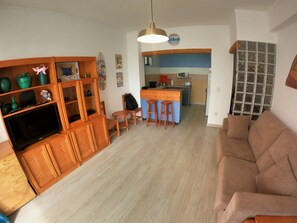 A living room with a sofa, a TV, wooden cabinets, and a kitchen area with a bar stool. - Algarve, modern T1 apartment with/balcony, Quarteira (Quarteira)