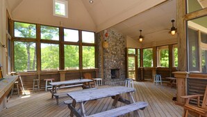 Un espacio de comedor al aire libre cubierto, con bancos y mesas de picnic de madera, ventanas amplias y una chimenea de piedra.
