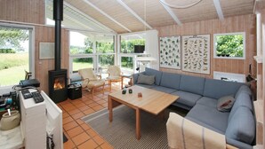 A cozy living room with a fireplace, a sofa, a coffee table, and a TV. There are framed pictures on the wall and a window with a view of greenery. - 2 bedroom lovely home in Samsø (Samsø)