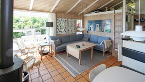 A cozy living room with a sofa, coffee table, and a fireplace. There are framed pictures on the wall and a window with a view of greenery outside. - 2 bedroom lovely home in Samsø (Samsø)