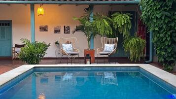 Une piscine agrémentée de deux fauteuils en osier et d’une petite table, le tout entouré de verdure et surmonté d’un toit en tuiles.