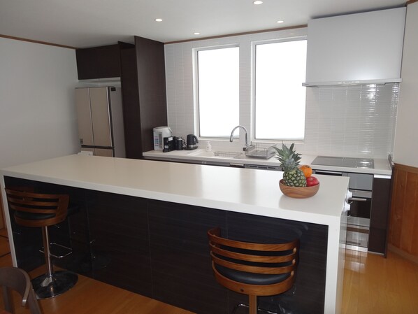 A modern kitchen with a central island, two bar stools, a refrigerator, a coffee maker, and a fruit bowl.