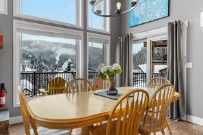 A dining area with a large table and chairs, a vase with flowers, and a view of a snowy landscape through large windows.
