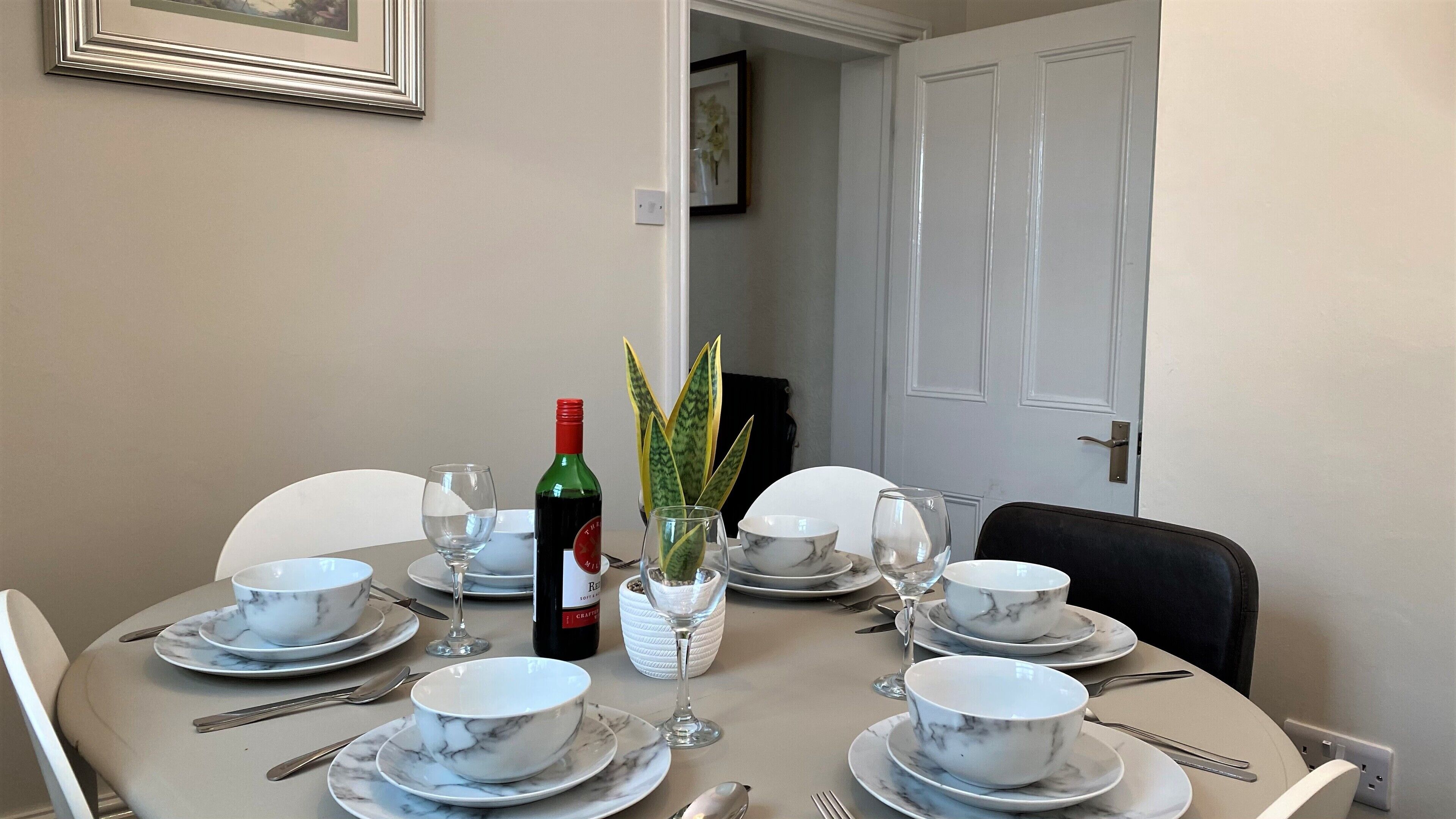 A dining table set with white marble plates, bowls, and wine glasses, with a bottle of red wine and a vase with green leaves.