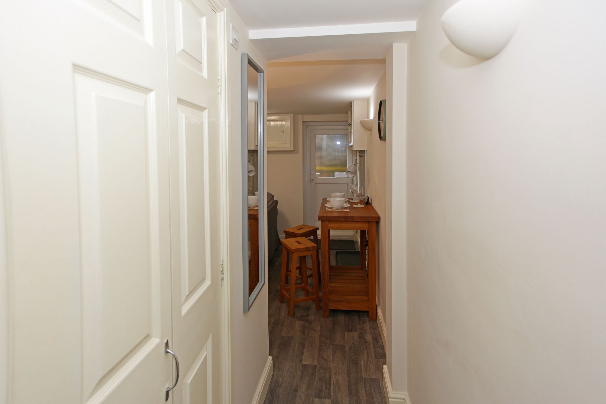 Passadís estret amb una taula i cadires de fusta, un mirall i una porta que dona a una altra habitació.