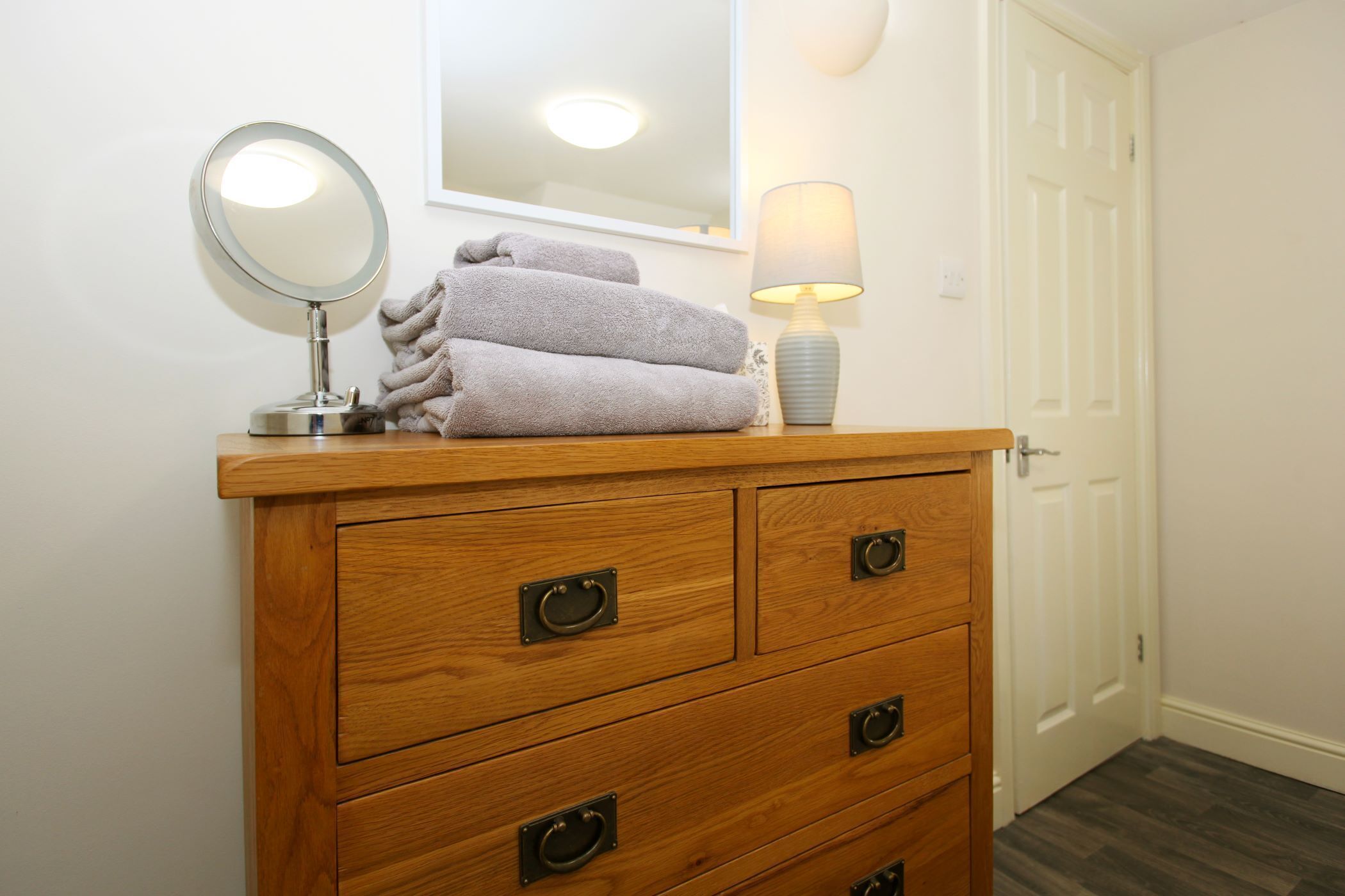 A wooden dresser with a round mirror, a lamp, and folded towels.