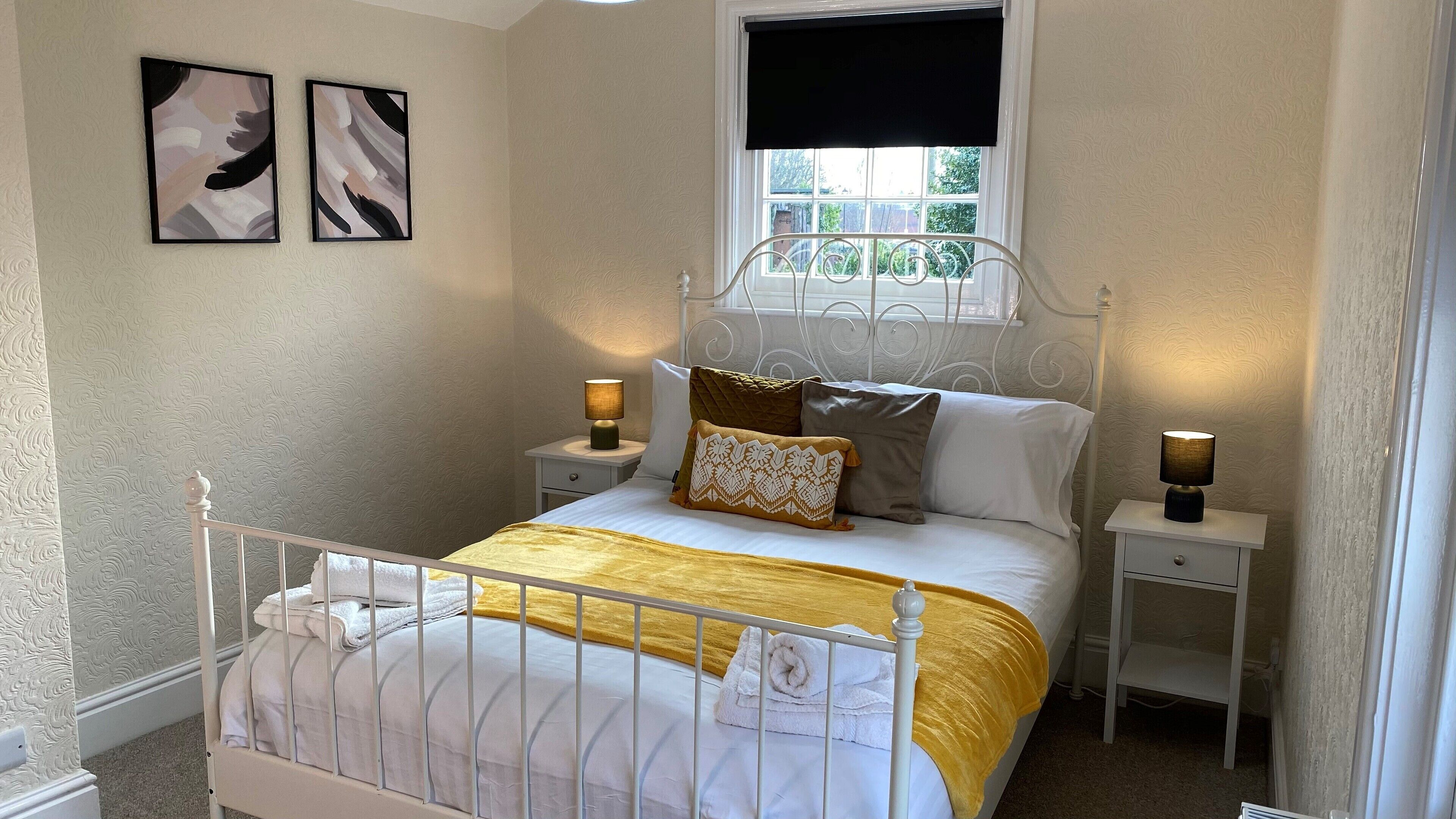 A bedroom with a bed, two bedside tables, a window, and two wall-mounted paintings.