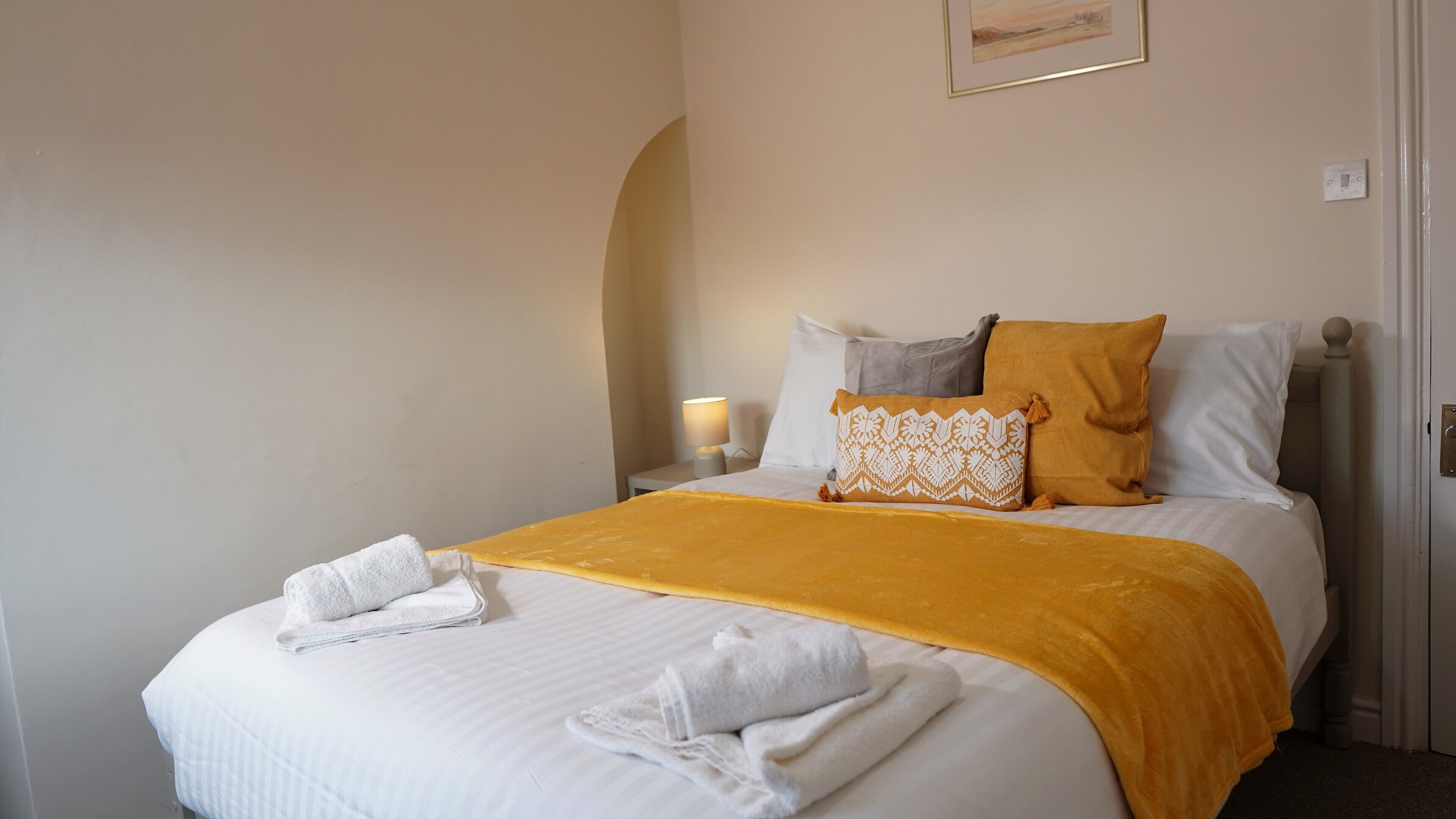 Una cama bien hecha con una colcha blanca, un edredón amarillo y varios almohadones, incluyendo uno estampado.