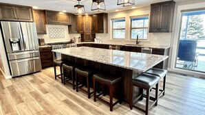 Una cocina moderna con una isla grande, armarios de madera, electrodomésticos de acero inoxidable y vistas al exterior a través de amplias ventanas.