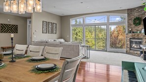 A spacious dining area with a wooden table set for four, surrounded by chairs. A large window offers a view of trees and a stone fireplace. - Welcome To Your New Favorite Spot! Tree Top Views & Lots Of Room! (Salem)