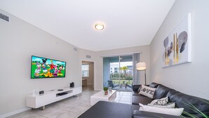 A modern living room with a flat-screen TV, a black leather sofa, a white entertainment unit, and a large window with a view of the outdoors.