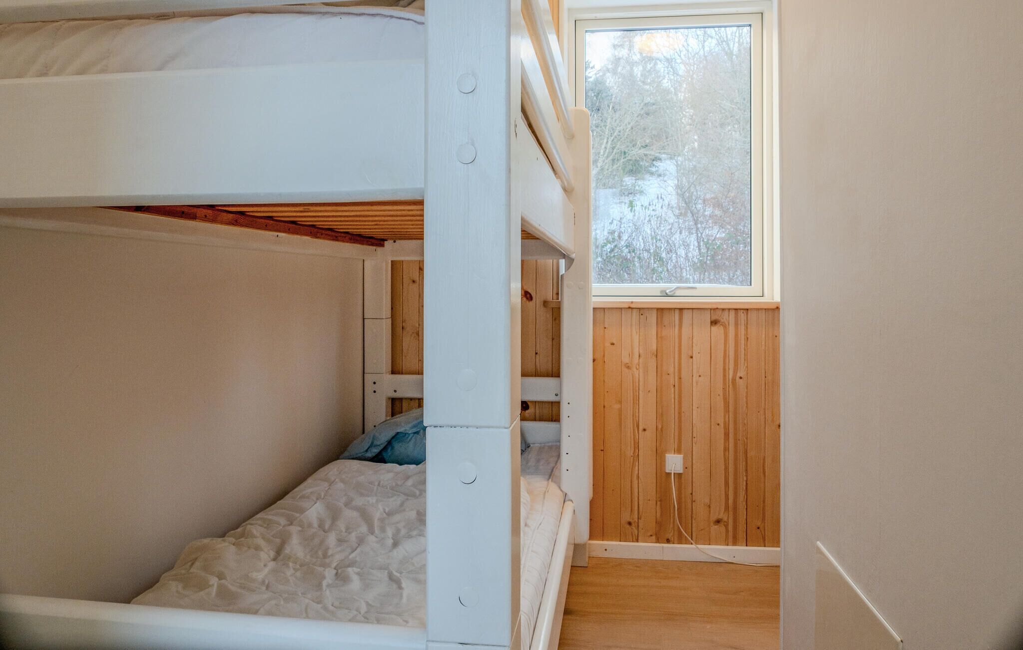 A small, single-room apartment with a bunk bed, wooden walls, a window, and a power outlet.