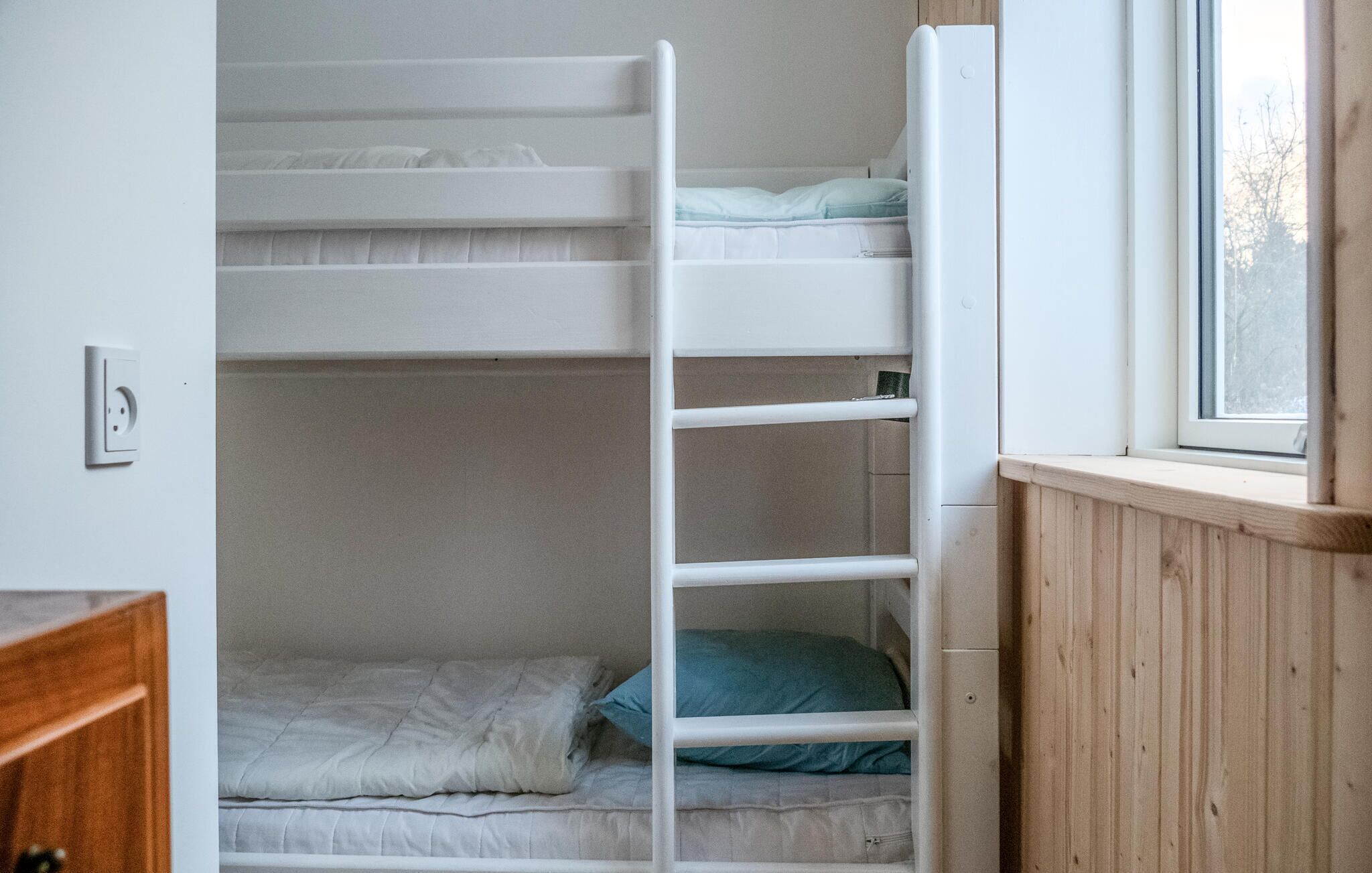 Une chambre avec un lit superposé, une fenêtre, une commode en bois et une prise électrique au mur.