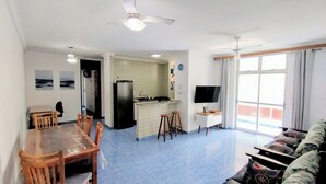 Ein Wohnzimmer mit einer Couch, einem Esstisch mit Stühlen, einer Küchenzeile inklusive Kühlschrank, einem Fernseher und einem Balkon mit Blick.