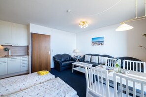 Una habitación de hotel compacta con kitchenette, dos camas, un sofá, una mesa de comedor y un plafón.