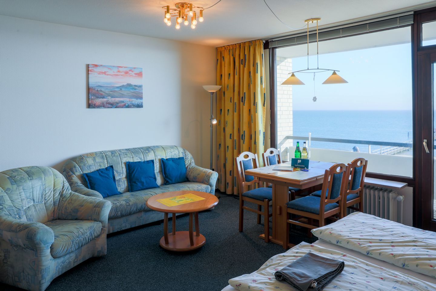A hotel room with a sofa, a small table with chairs, a bed, and a view of the sea through a large window.