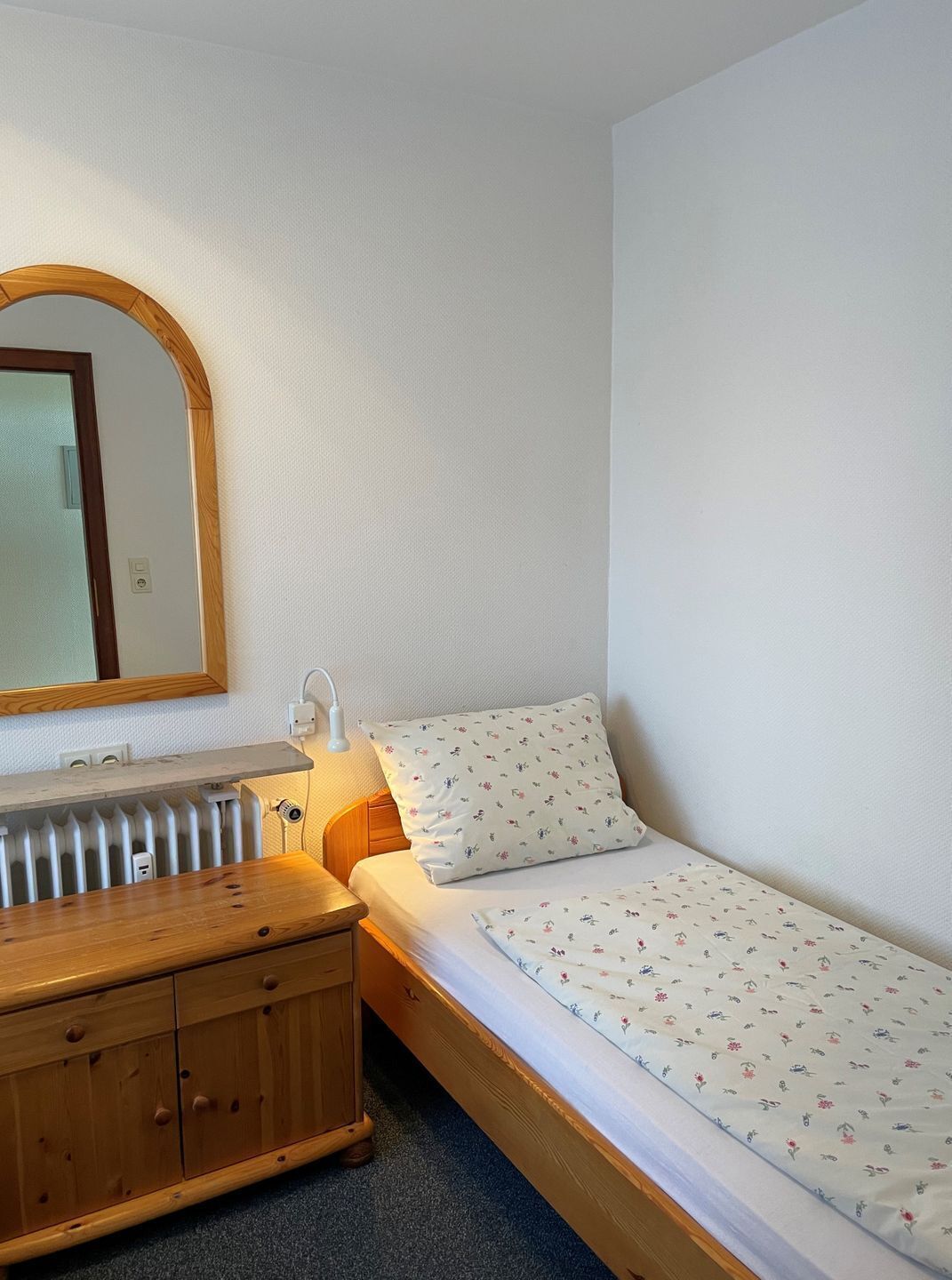 A small, single-bed room with a wooden dresser, a mirror, and a radiator.