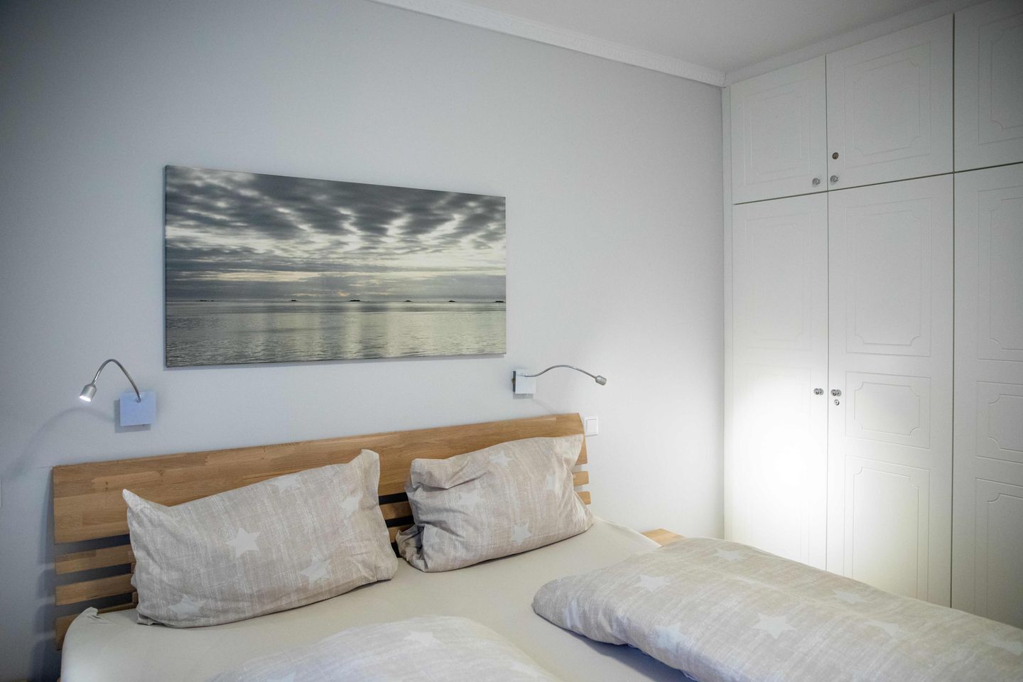 Un dormitorio con cabecera de madera, dos mesitas de noche con lámparas, un cuadro grande de un atardecer y un armario con puertas.