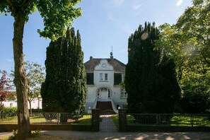 A white villa with a central entrance, surrounded by tall trees and a metal fence.