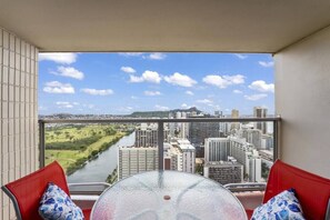 A balcony with a glass table and red chairs, overlooking a cityscape and a river.