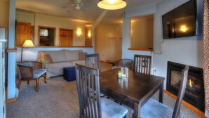 A cozy living room with a fireplace, a dining table, and a TV mounted on the wall. - 7 Grandview - Unit D - Great condo close to downtown Rangeley (Rangeley)