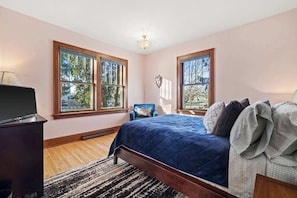 Une chambre à coucher avec un lit, deux fenêtres, une chaise, un bureau et une lampe.