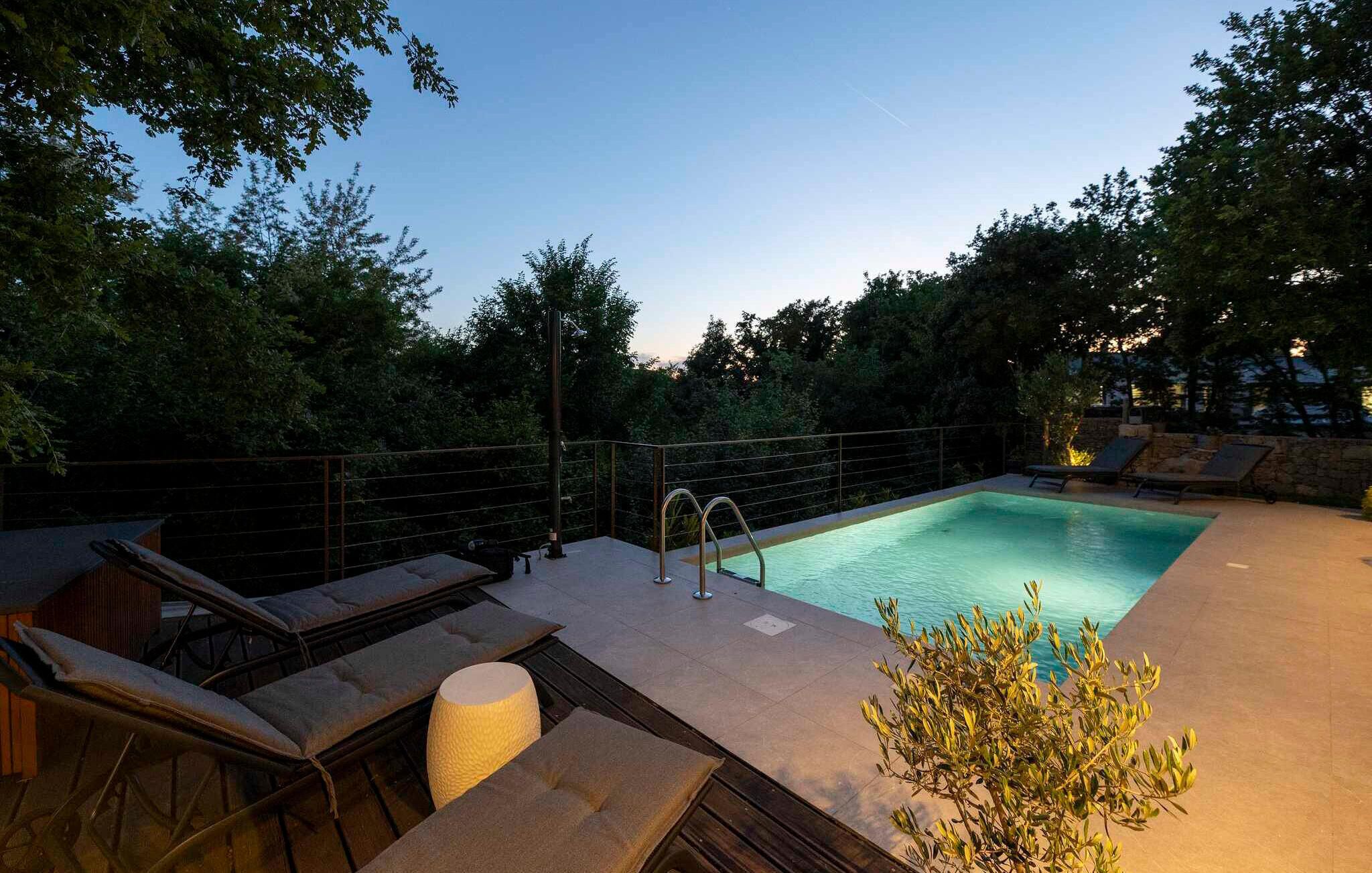 Una piscina rettangolare con una scala in metallo, circondata da un muro in pietra e alberi. Sono presenti sedie a sdraio e un tavolino con cuscini.