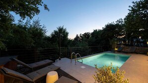 Une piscine rectangulaire avec un escalier en métal, entourée d’un mur de pierre et d’arbres. On y trouve des chaises longues et une petite table avec des coussins.