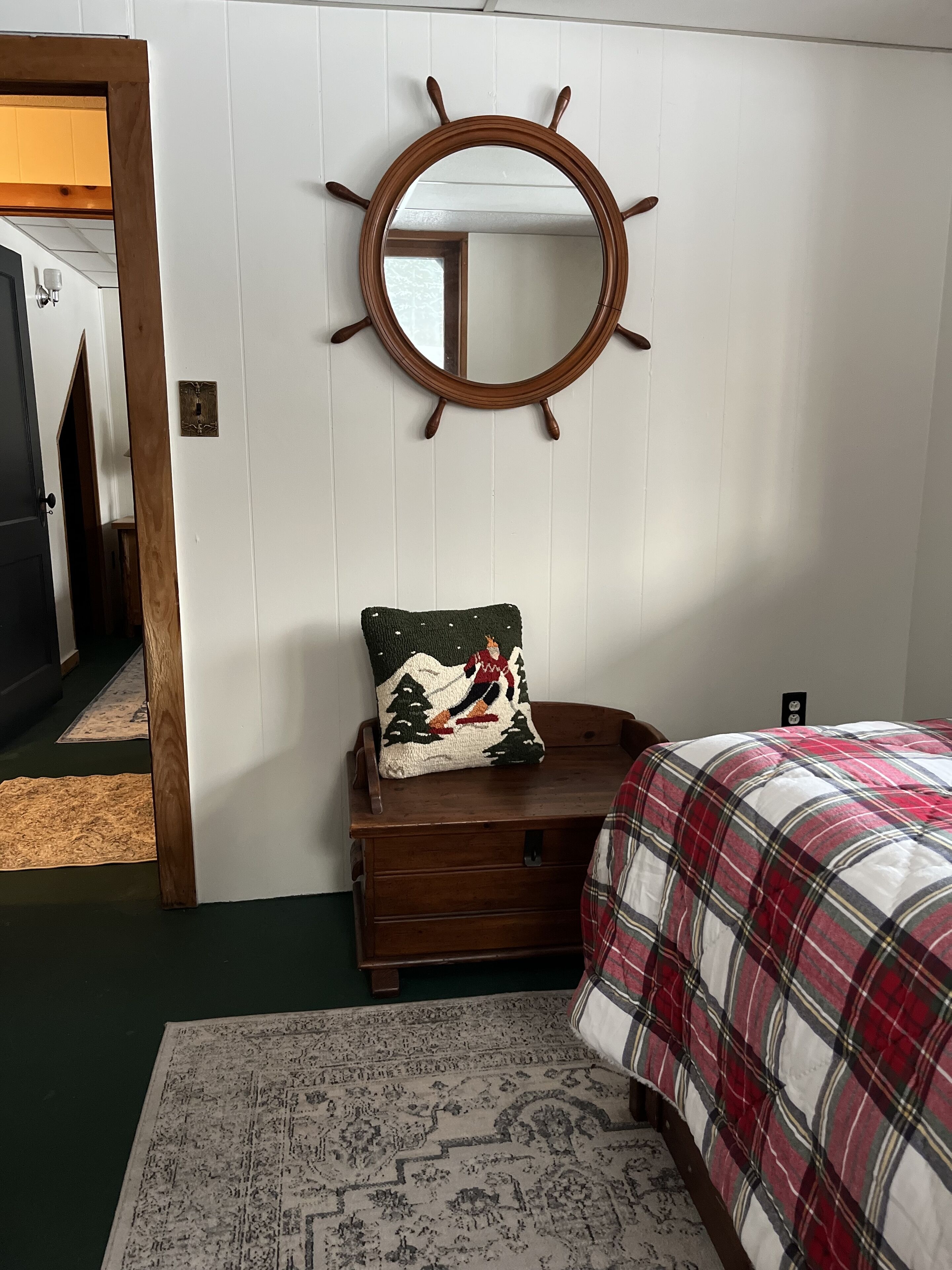 A bedroom with a bed, a dresser, a mirror with a ship's wheel design, a decorative pillow, and a patterned rug.