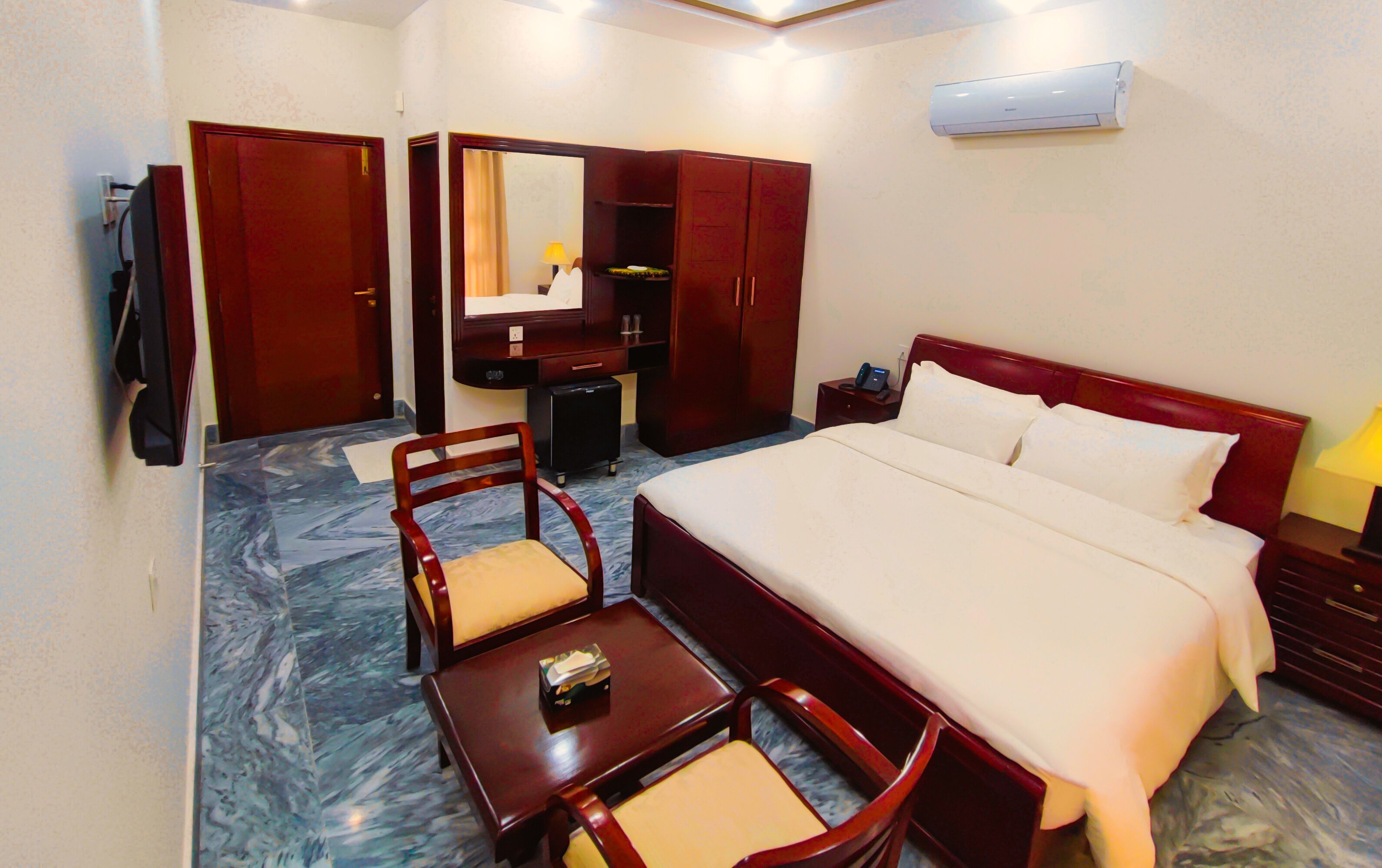 A hotel room with a bed, two chairs, a desk, a wardrobe, a mirror, a bedside table, and an air conditioner.