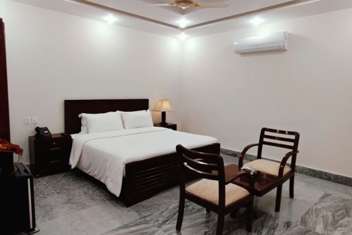 A bedroom with a bed, bedside table, lamp, chair, and an air conditioning unit.