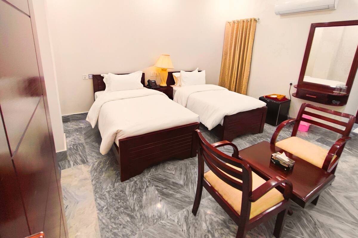 A hotel room with two beds, a desk with a chair, a mirror, a lamp, and a small table.