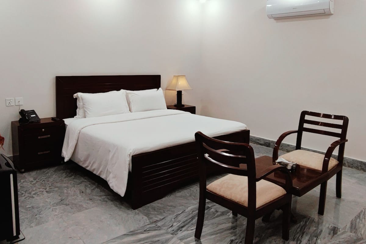 A hotel room with a bed, bedside table with a lamp, a chair, a desk with a computer, and a water bottle.