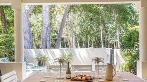 An outdoor dining area with a wooden table set for four, surrounded by white chairs. There are potted plants, a wine bottle, and a fruit platter on the table. - Sea La Vie - Port Douglas Villa (Port Douglas)
