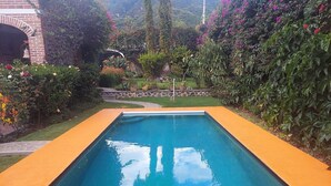 A rectangular swimming pool with a smooth, reflective surface, surrounded by a low brick wall and a variety of lush greenery and flowering plants. - Casita. W PRIVATE POOL (Ajijic)