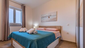 A neatly made bed with a teal bedspread and pink pillows, a painting of a coastal scene on the wall, and a window with curtains.