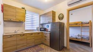 A compact kitchen with wooden cabinets, a stainless steel refrigerator, a microwave, and a coffee maker. There's a window with blinds, a clock on the wall, and a bunk bed with a wooden ladder.