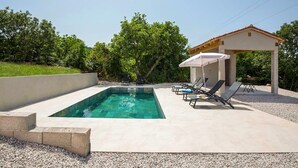 A rectangular swimming pool with a concrete deck, surrounded by lounge chairs and a covered seating area.