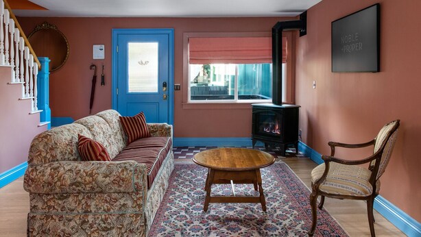 A living room with a floral sofa, a round wooden coffee table, a patterned rug, a blue door, a black wood-burning stove, and a chair with a striped cushion.