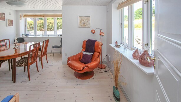 Quarto com mesa de jantar, cadeiras, poltrona de couro, luminária de chão e janela com vista para vegetação.