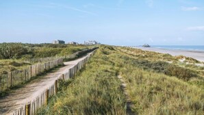 Een kustpad met een houten omheining, grasduinen en gebouwen in de verte.