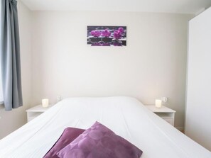 Un lit bien fait, avec du linge de lit blanc et deux coussins violets, une petite table de chevet avec une lampe blanche et un tableau représentant des fleurs violettes accroché au mur.